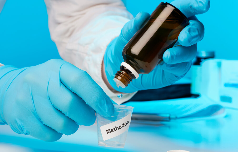 Arzt mit blauen Handschuhen Tropft Methadon in einem Kleinen Becher. Das Ettikett auf dem Becher ist Englisch. Der Hintergrund ist blau Der Becher ist auf deutsch beschriftet